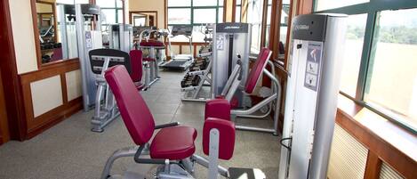 Un gimnasio con diferentes máquinas de ejercicio, ventanas amplias y piso de madera.