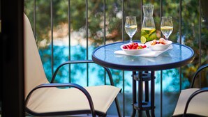 Une table ronde avec deux chaises, un saladier de fruits et deux verres de vin blanc, le tout sur un balcon donnant sur une piscine.
