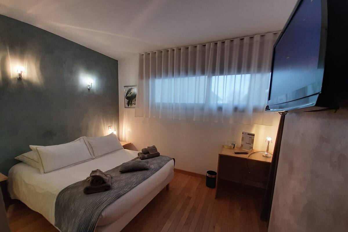 A modern hotel room with a bed, bedside tables, a TV, and a window with curtains.