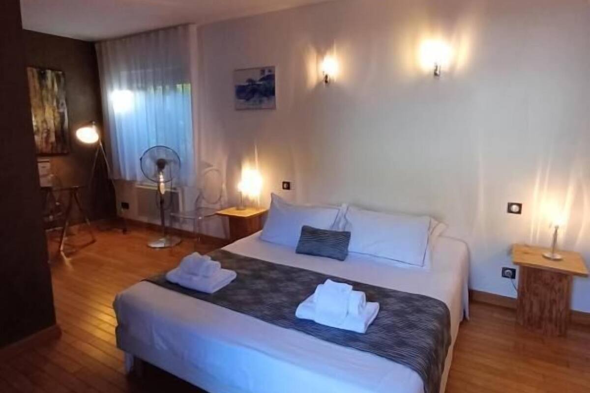 A hotel room with a large bed, two bedside tables, a standing fan, and wall-mounted lights.