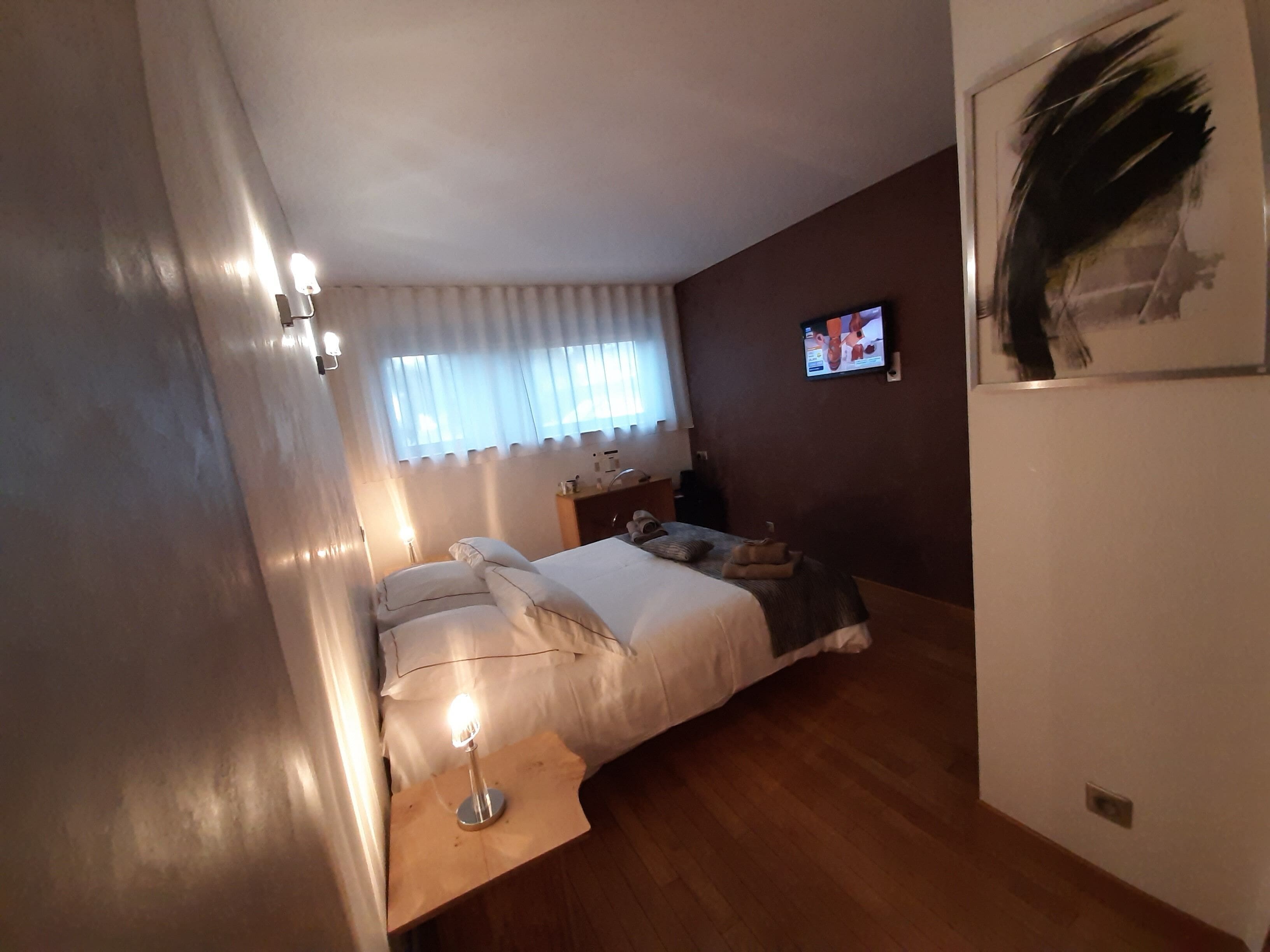 A bedroom with a bed, bedside lamps, a window with curtains, a TV on the wall, and a painting.
