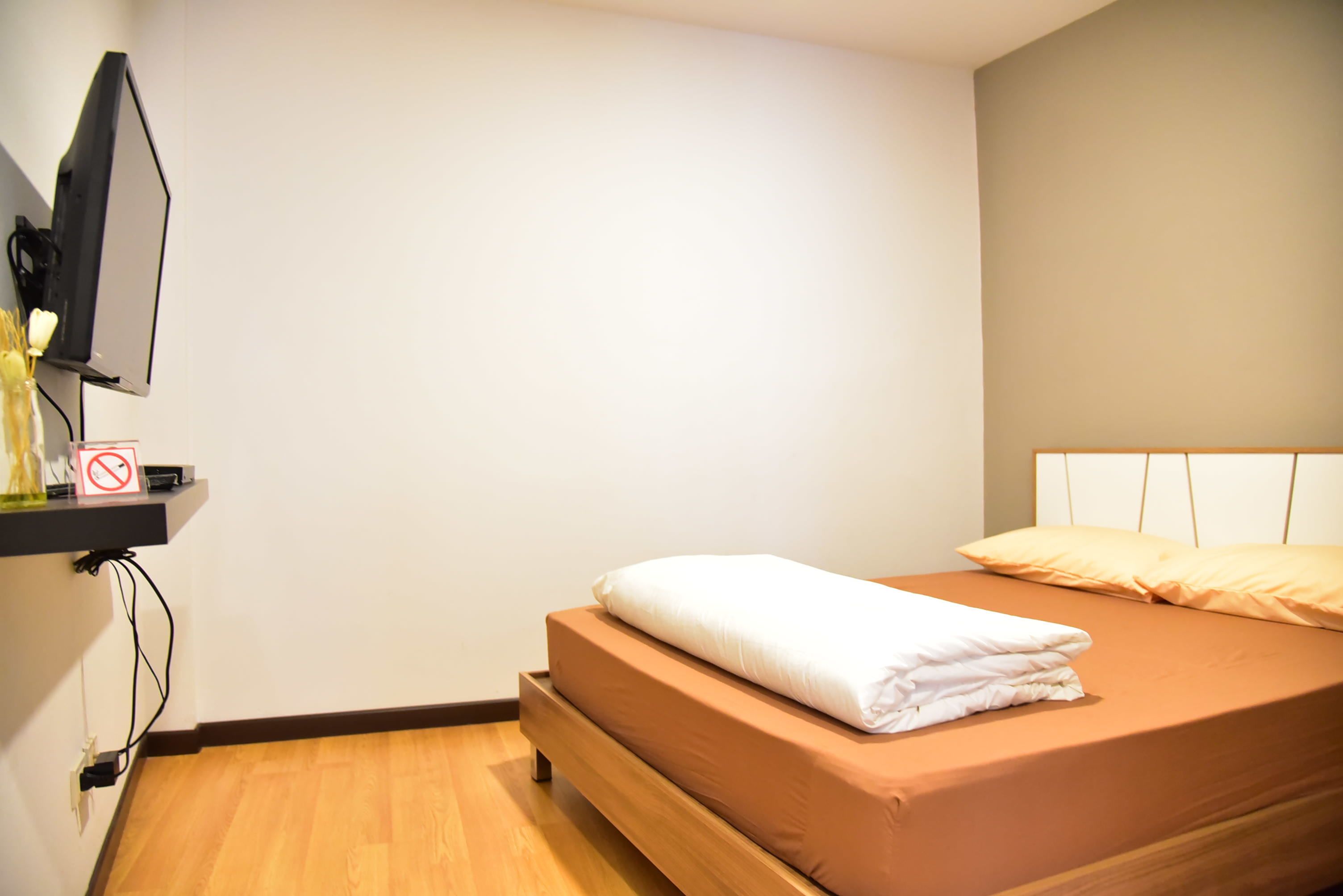A neatly made bed with a brown mattress and white bedding, a television mounted on the wall, and a small shelf with a no-smoking sign.