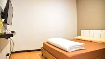 A neatly made bed with a brown mattress and white bedding, a television mounted on the wall, and a small shelf with a no-smoking sign.