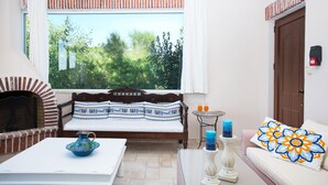 A cozy room with a brick fireplace, a sofa with patterned cushions, a small table with a pitcher, and a window with a view of greenery. - Villa Dafni (Faistos)