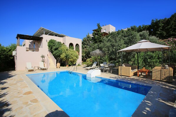 A swimming pool with lounge chairs and a covered seating area, surrounded by a landscaped garden and a building with a modern design. - Villa Dafni (Faistos)