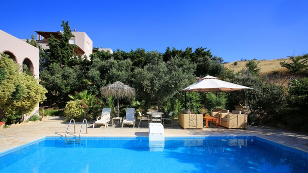 A swimming pool with lounge chairs and a covered area, surrounded by greenery and a building. - Villa Dafni (Faistos)