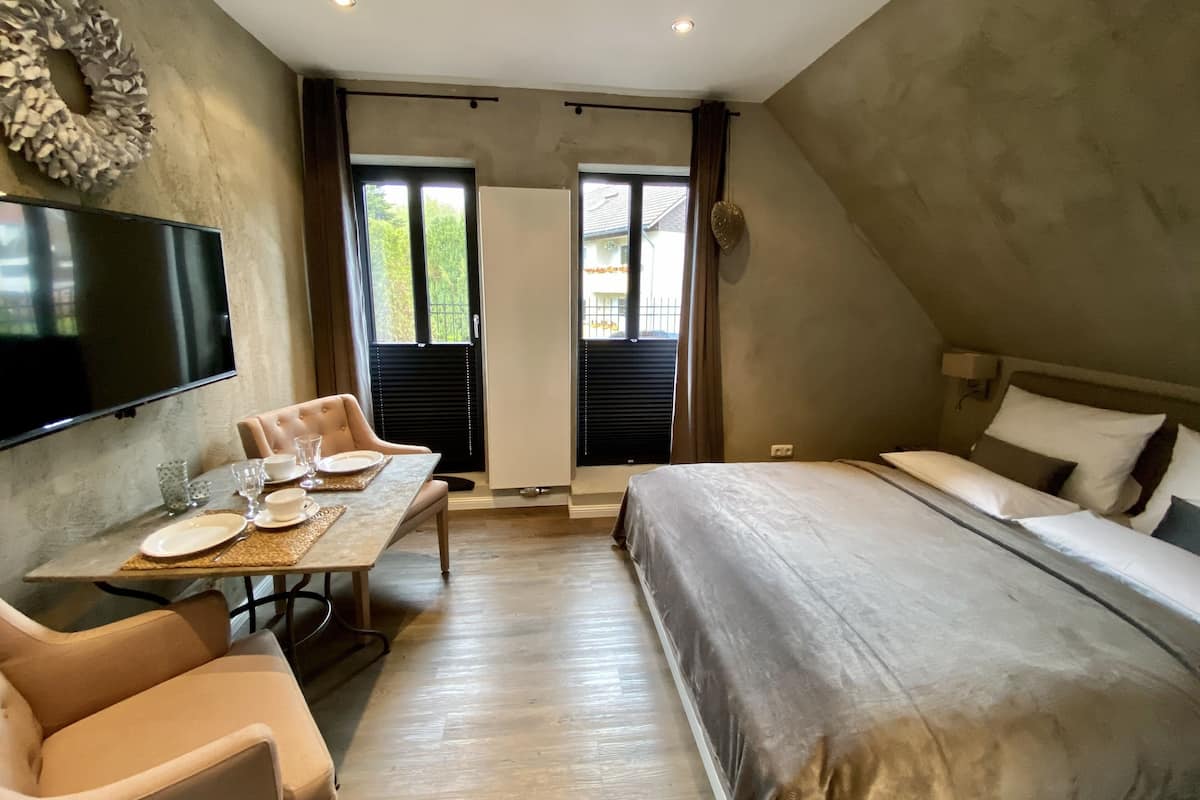 A bedroom with a bed, a TV, a dining table with chairs, and a view of the outdoors through a door.