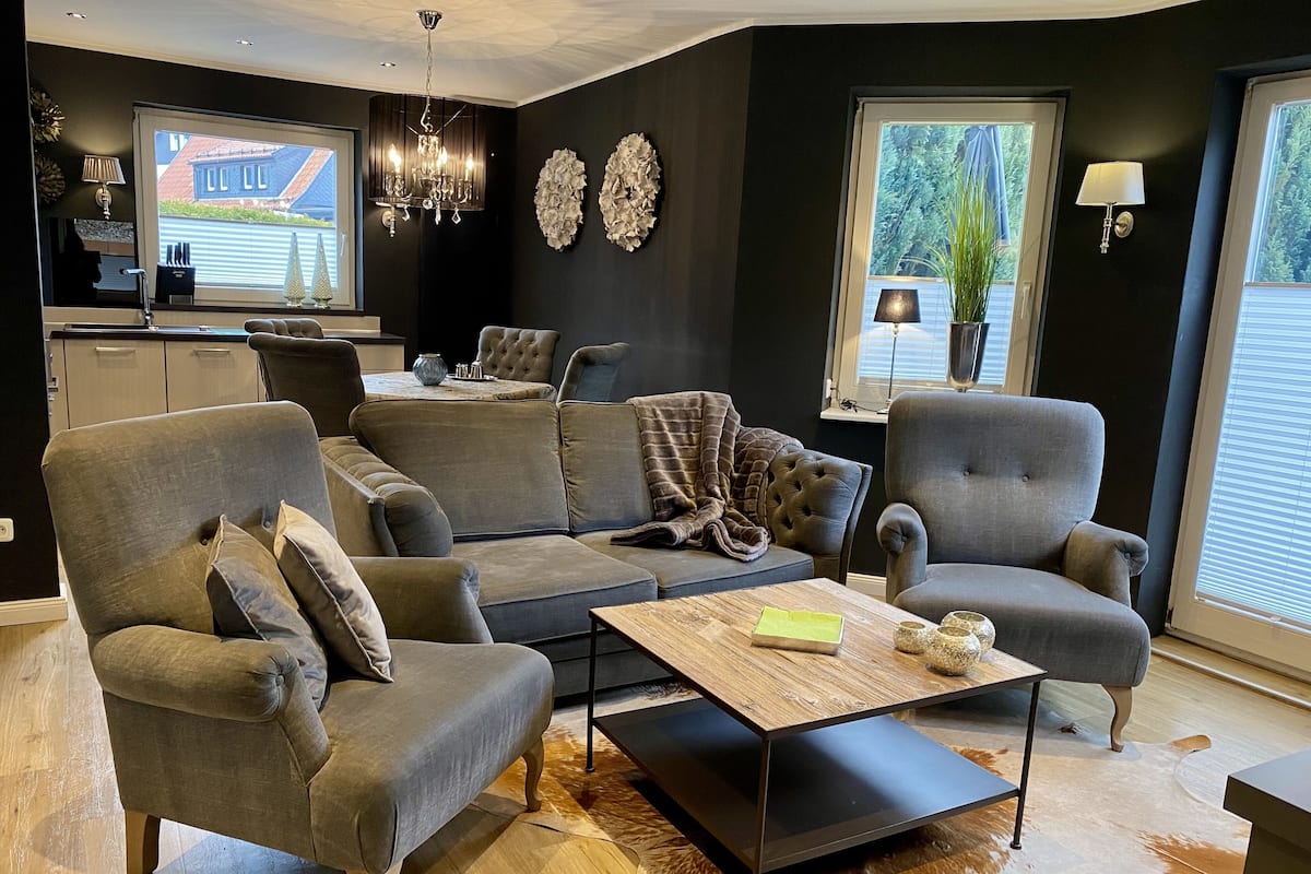 A modern living room with a grey sofa, armchairs, a coffee table, and a kitchen in the background.