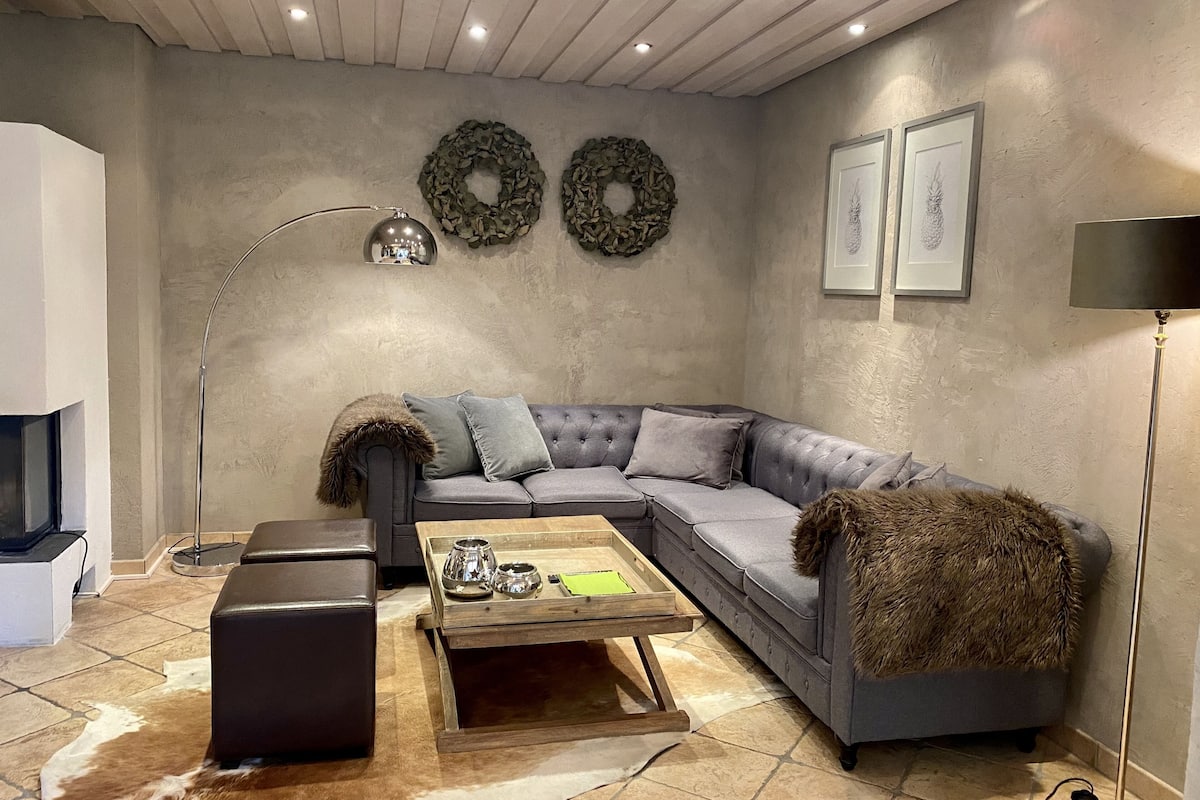 A modern living room with a grey sectional sofa, a wooden coffee table, a black ottoman, and a stone floor.