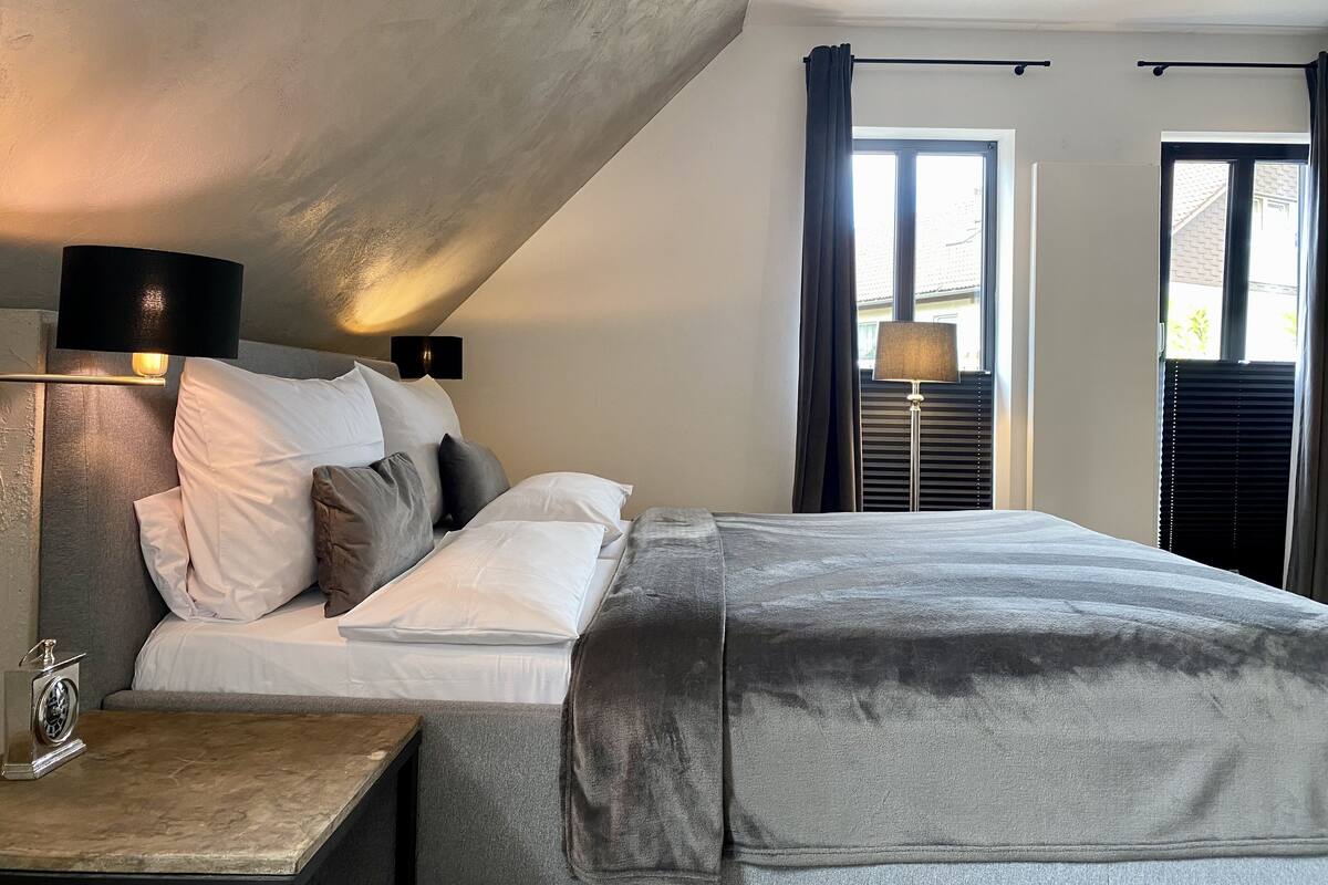 A modern bedroom with a large bed, two bedside lamps, a nightstand, and a window with curtains.