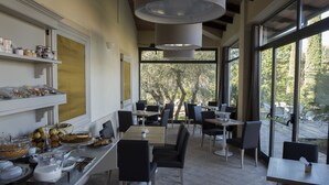Uma área de refeições com mesas e cadeiras, um buffet com diversos alimentos e grandes janelas com vista para árvores.
