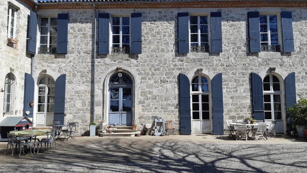 A stone building with arched windows and shutters, outdoor seating, and a gravel courtyard.