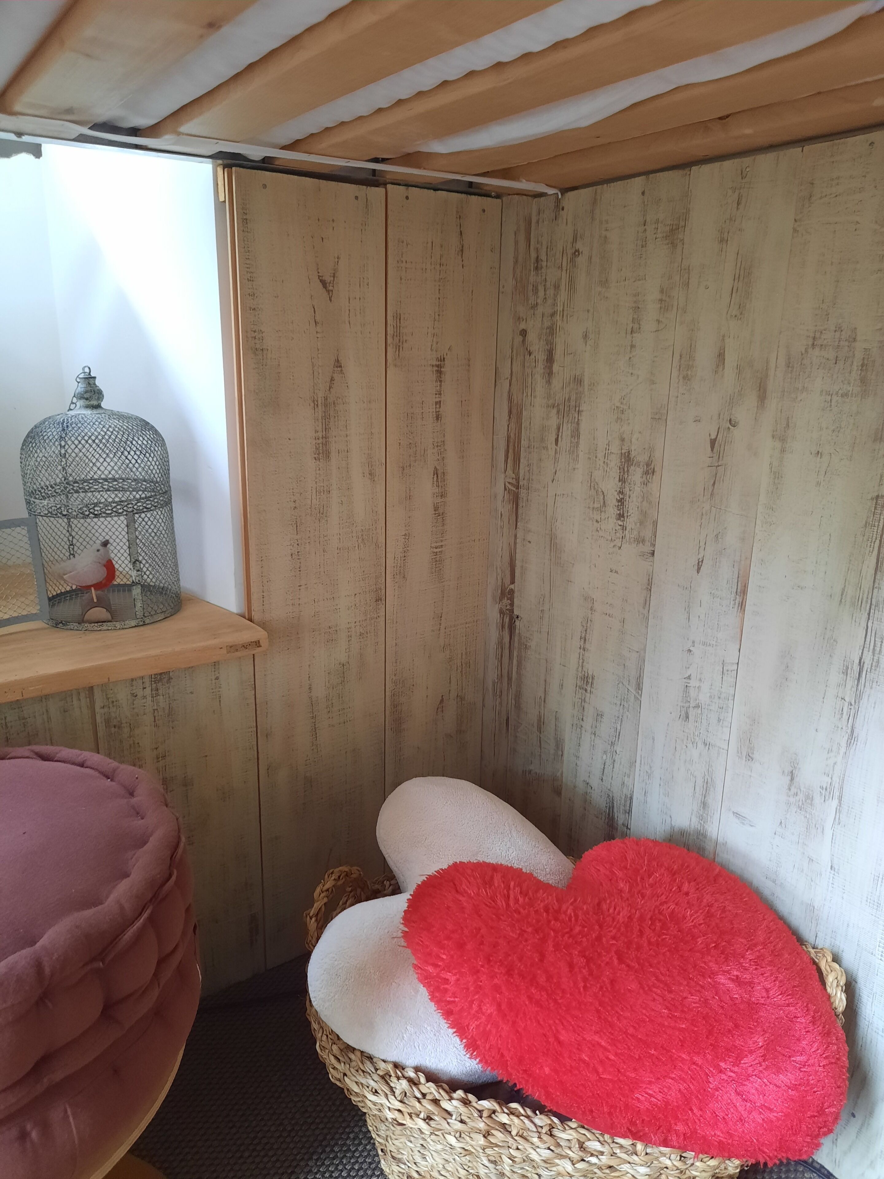 Canto de um quarto com parede de madeira, um cesto de vime com almofadas e uma gaiola de metal com um pássaro.