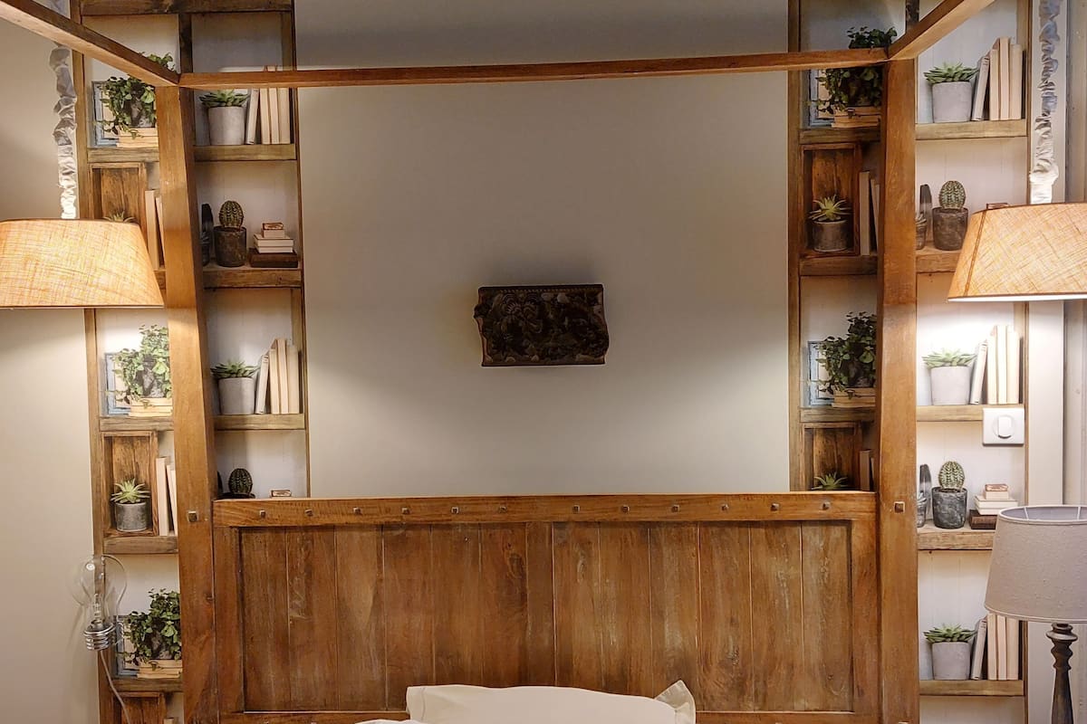 A bedroom with a wooden headboard, a bed with white bedding, and shelves with books and plants.