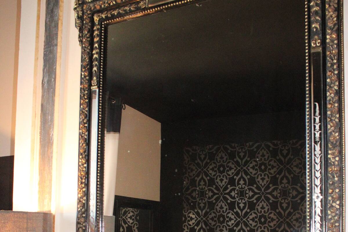 A bedroom with a large ornate mirror, a patterned wallpaper, a radiator, and a bedside table with a lamp.