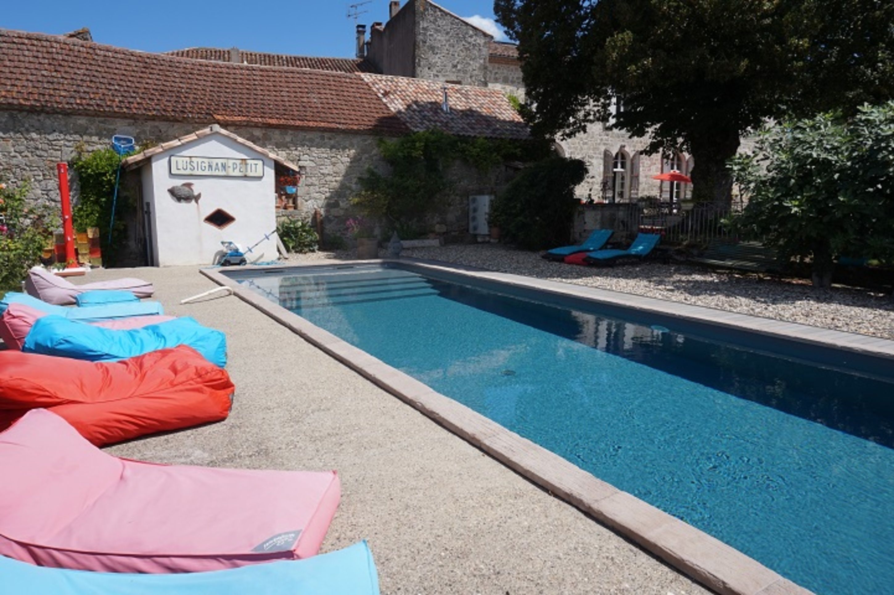 Piscina com espreguiçadeiras, um pequeno edifício com a placa 'Lusignan-Petit' e edifícios de pedra ao fundo.
