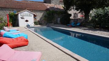 Piscina com espreguiçadeiras, um pequeno edifício com a placa 'Lusignan-Petit' e edifícios de pedra ao fundo.