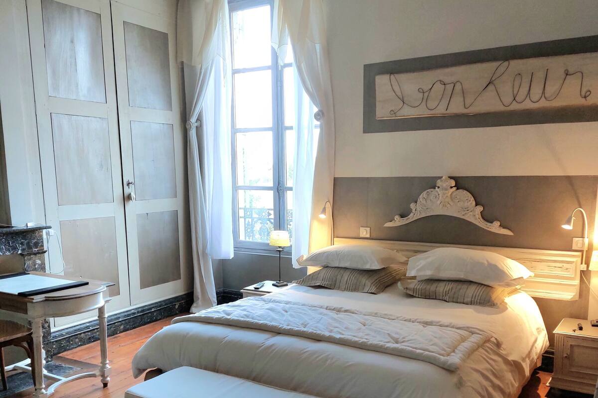 A bedroom with a large bed, a bedside table, a nightstand, a bench, and a window with curtains.