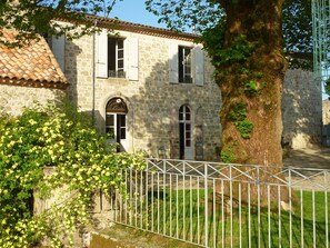 A stone building with a tiled roof, a tree with green foliage, and a metal fence. - Au Bonheur d'Alphonse (Lusignan-Petit)