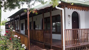 A traditional wooden porch with a brick pathway leading to a white building with a dark door. - Green Olive Guesthouse (Robertson)