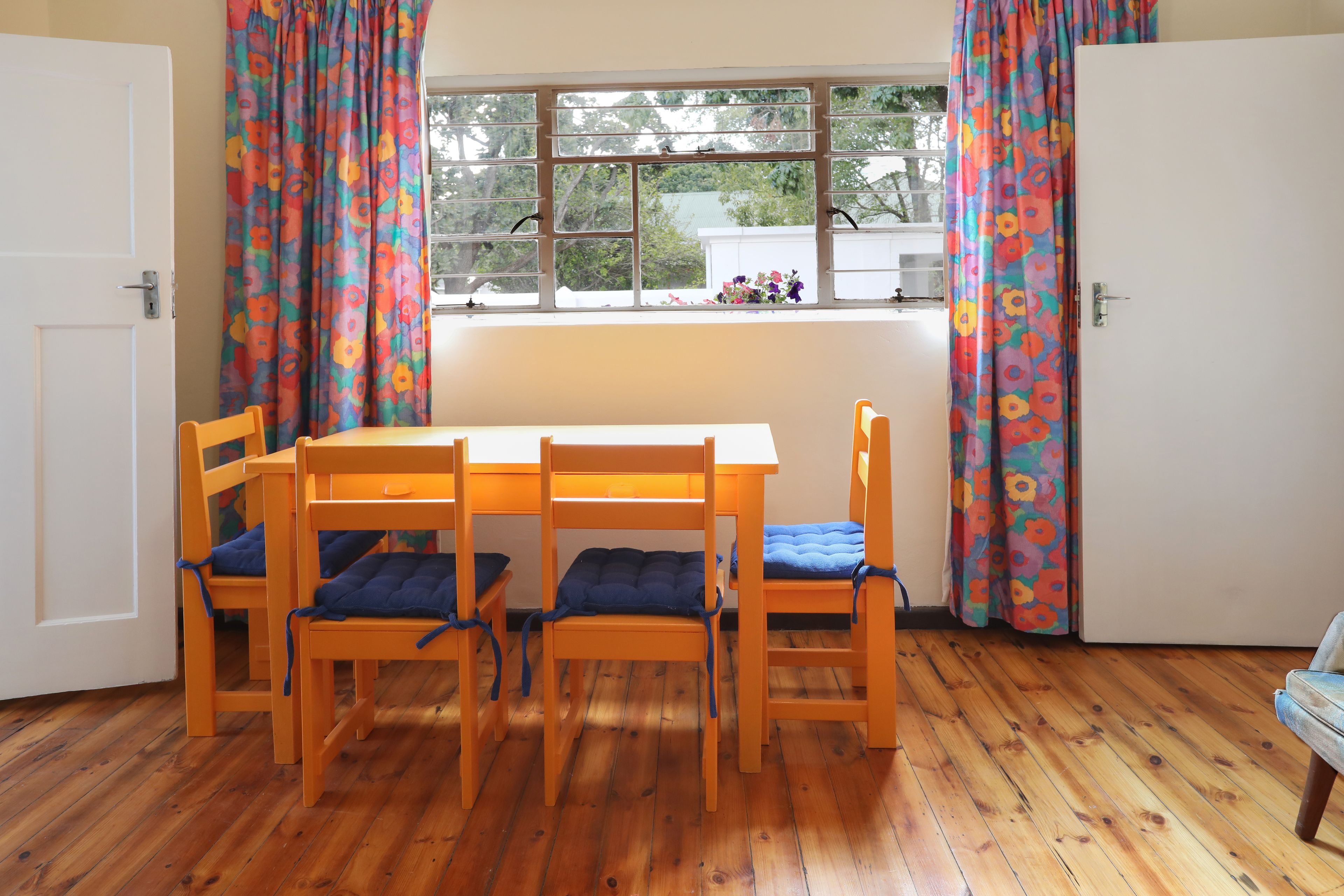 A room with a wooden table and four chairs, a window with curtains, and a door.