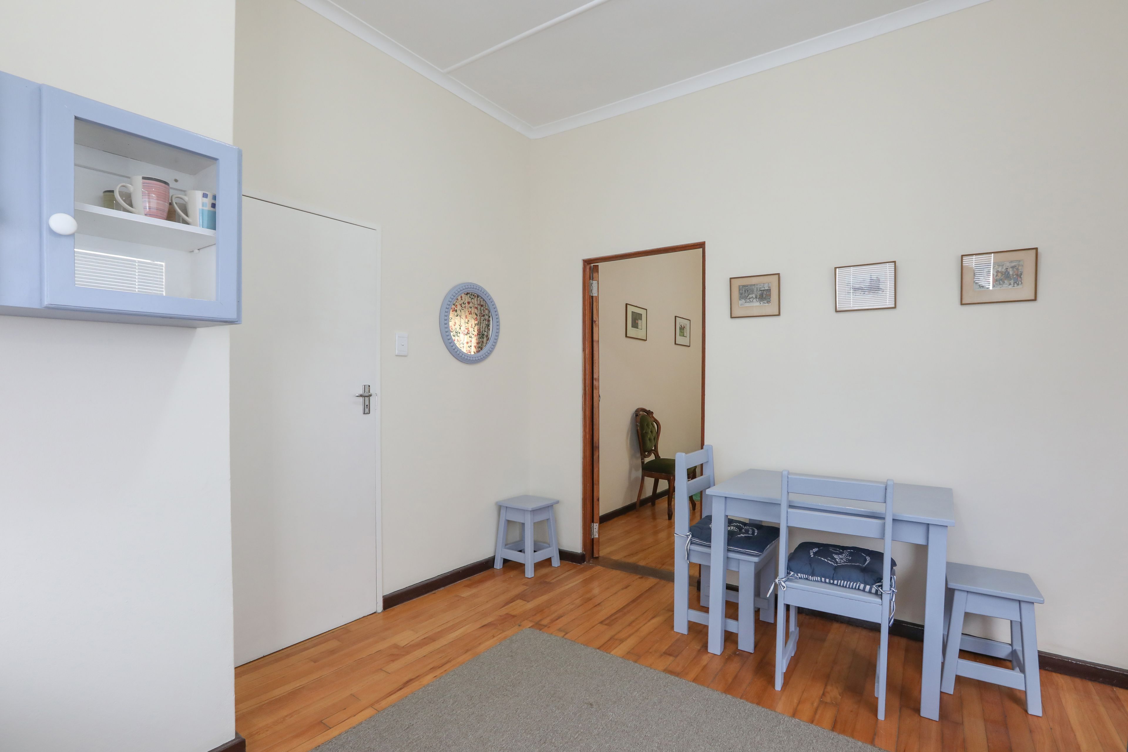 Una habitación con piso de madera, un escritorio con sillas, un taburete pequeño, un mueble con tazas y cuadros en la pared.