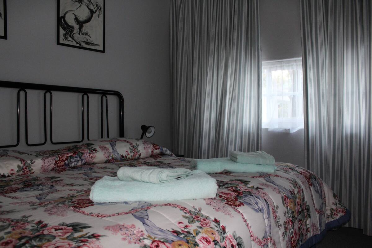 Ein Schlafzimmer mit floralem Bettbezug, einem ordentlich bezogenen Bett mit gefalteten Handtüchern, einem schwarzen Metall-Kopfteil, einem Fenster mit Vorhängen und einem gerahmten Bild an der Wand.