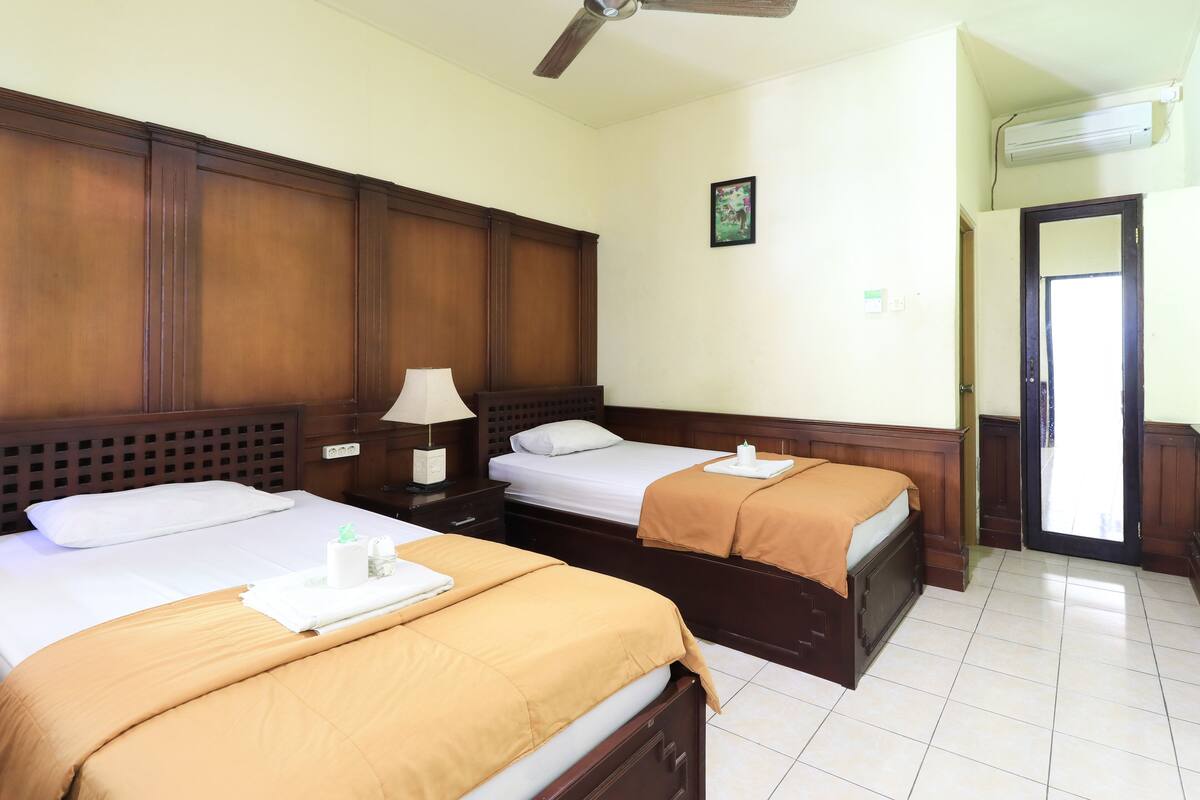 Habitación de hotel con dos camas, cabeceros de madera, ventilador de techo, mesita de noche con lámpara y puerta que da a un balcón.