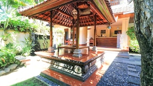 A traditional wooden gazebo with a tiled floor, surrounded by a garden with various plants and a stone pathway. - Sari Indah Cottages (Kuta)