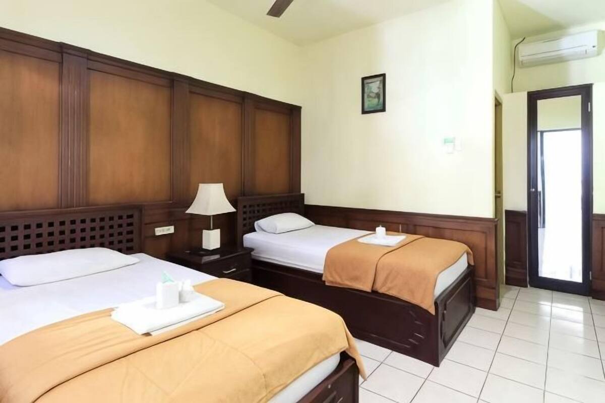 Habitación de hotel con dos camas, una mesita de noche con lámpara, cabecera de madera, piso de baldosas y una puerta con equipo de aire acondicionado.
