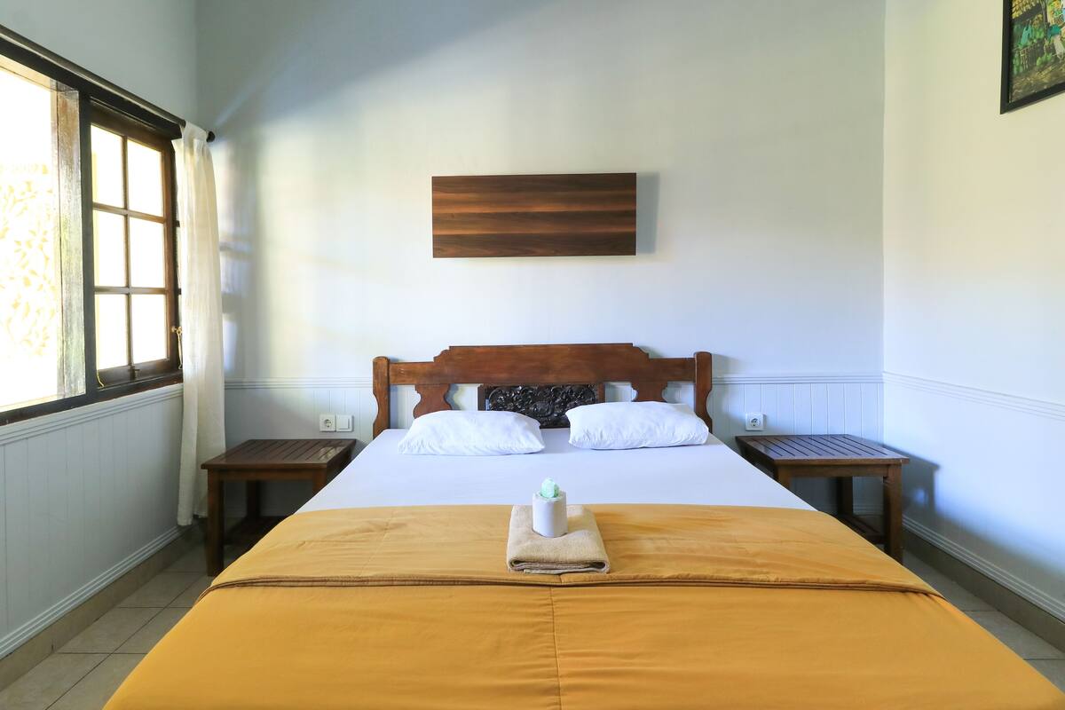 Una cama cuidadosamente tendida con una colcha marrón, almohadas blancas y una pequeña jarra blanca en el centro. Se aprecia un cabecero de madera, dos mesitas de noche y una obra de arte colgada en la pared sobre la cama.