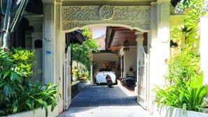 A narrow pathway leading to a building with ornate architectural details, a white car parked in front, and potted plants on either side. - Sari Indah Cottages (Kuta)