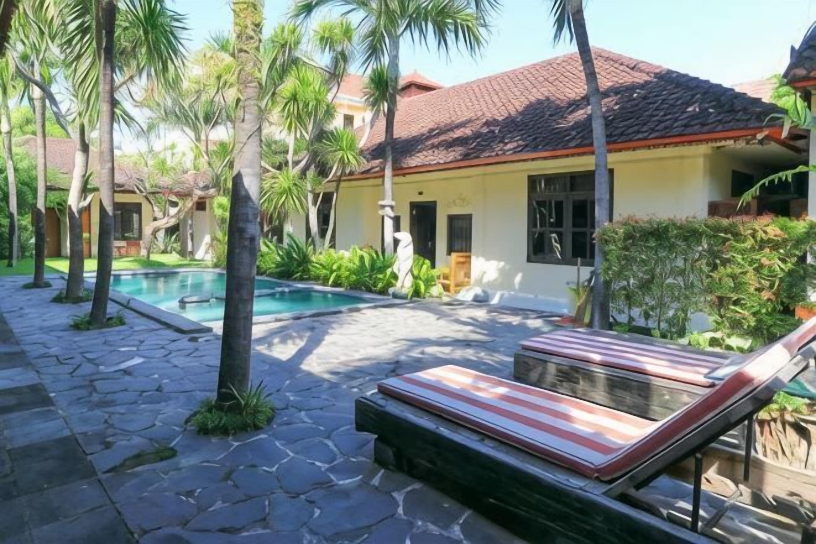A pool area with a lounge chair, a stone pathway, and a building with a red-tiled roof.