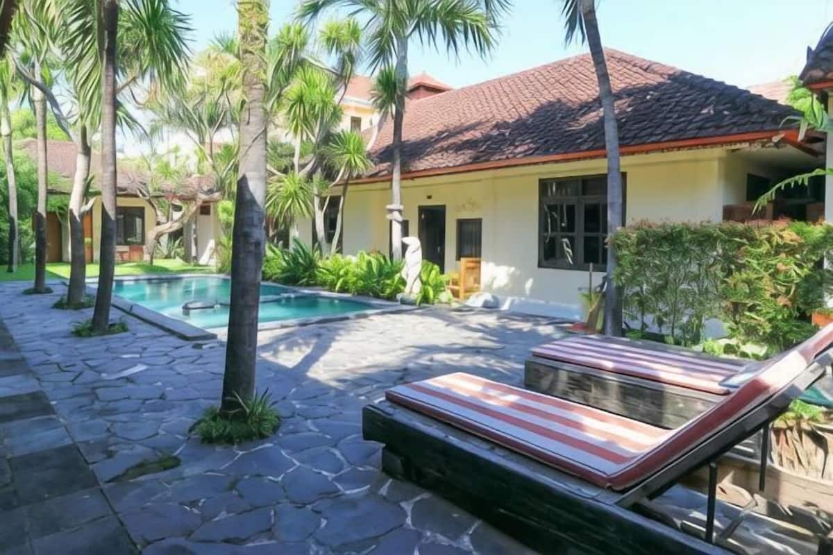 A pool area with a lounge chair, a stone pathway, and a building with a red-tiled roof.