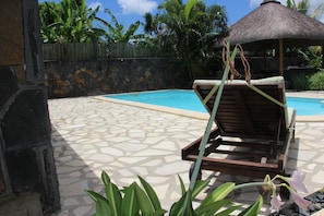 A pool area with a lounge chair, a thatched roof structure, and a stone wall.