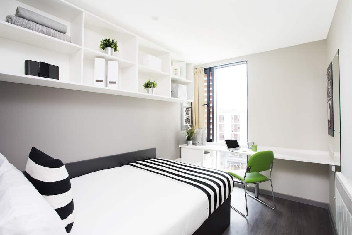 A modern bedroom with a bed, a desk, and a chair. There is a window with a view of buildings outside.