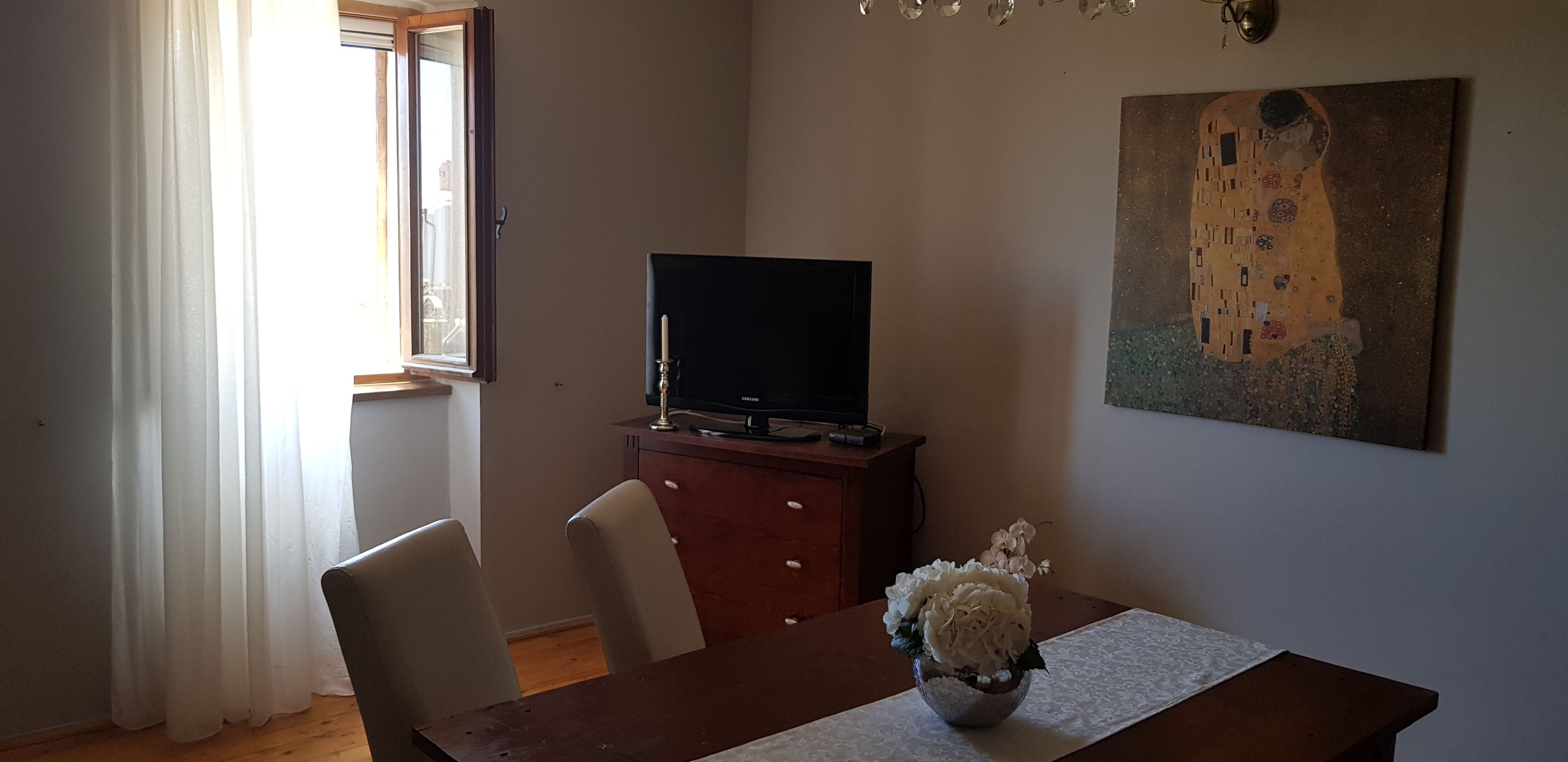 Een woonkamer met een houten tafel, een televisie op een kast, een schilderij aan de muur en een raam met gordijnen.