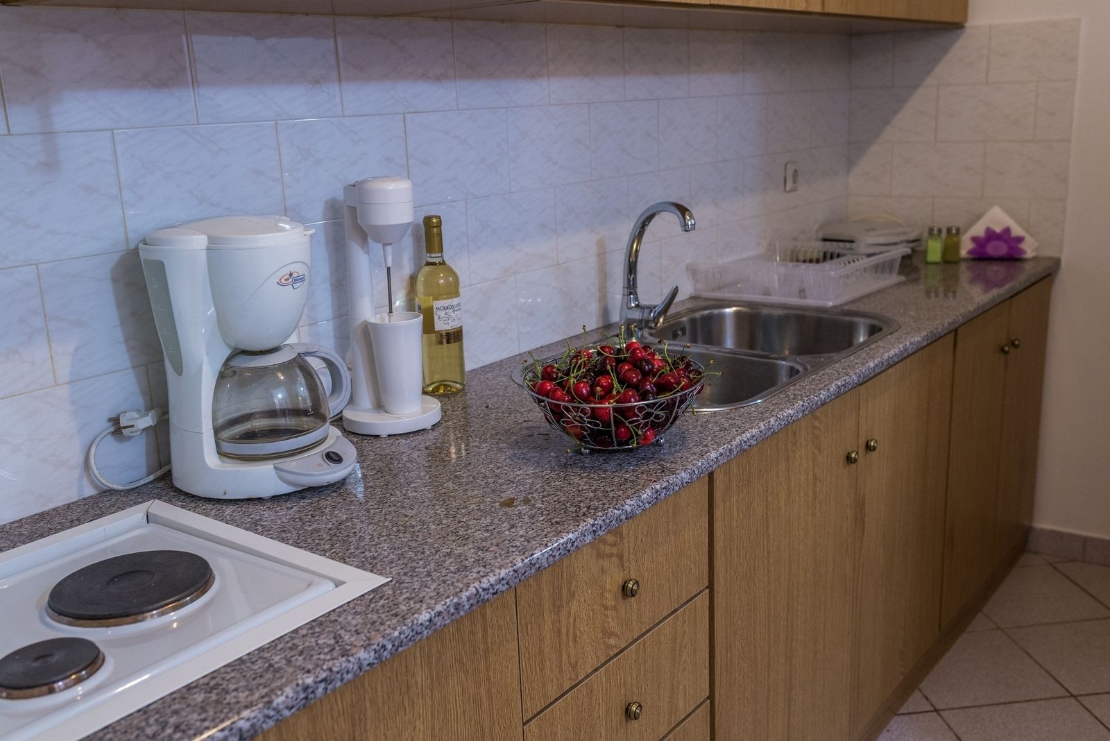 Una cocina con cafetera, botella de vino, un bol de cerezas y un lavabo.