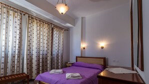 Un dormitorio con cama, cabecera de madera, dos mesitas de noche, un banco y cortinas.