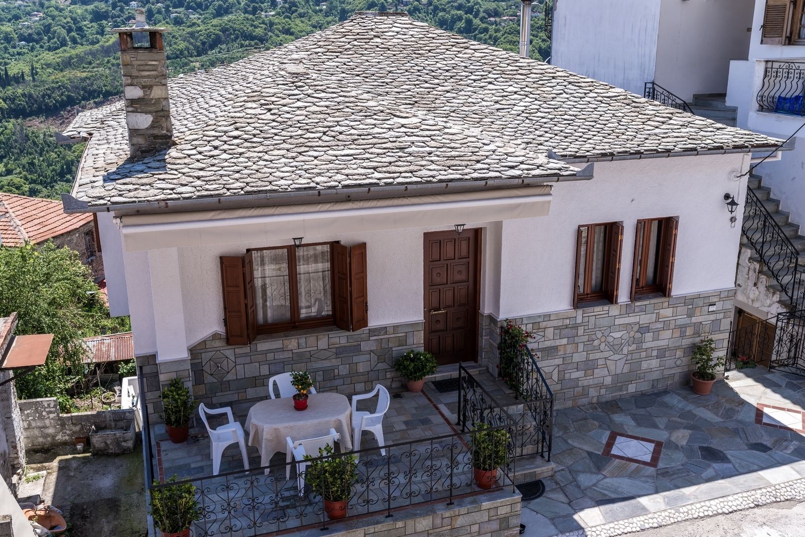 A two-story house with a stone roof, a balcony with a table and chairs, and a garden area.