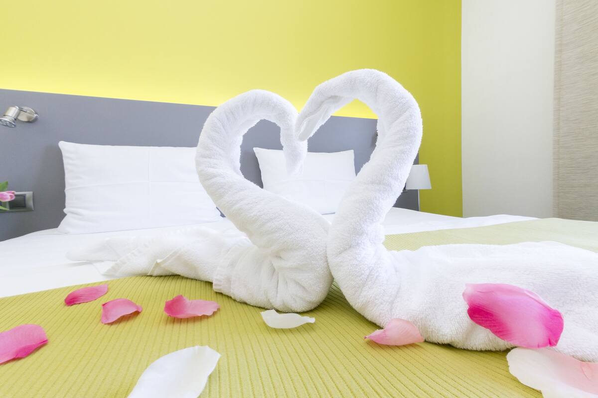 Una habitación de hotel con una cama, almohadas, una mesita de noche y una lámpara de pared.