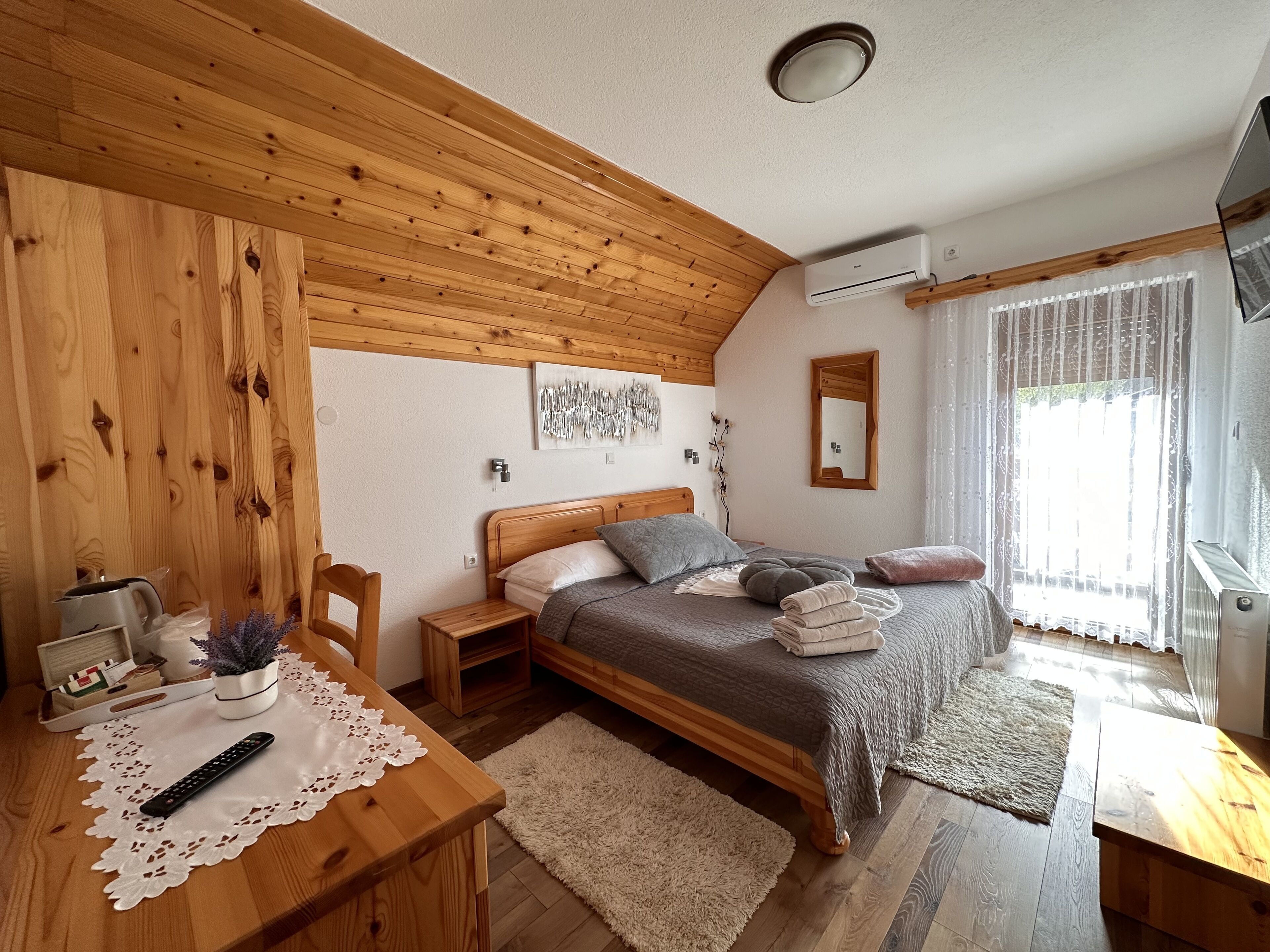 Un dormitorio acogedor con techo de madera, una cama con manta gris, una mesita de noche con hervidor y una ventana con cortinas translúcidas.
