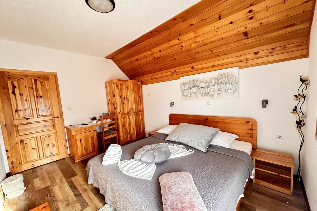 Un dormitorio con techo de madera, una cama con almohadas y manta, un armario de madera, un escritorio con silla y una mesita.