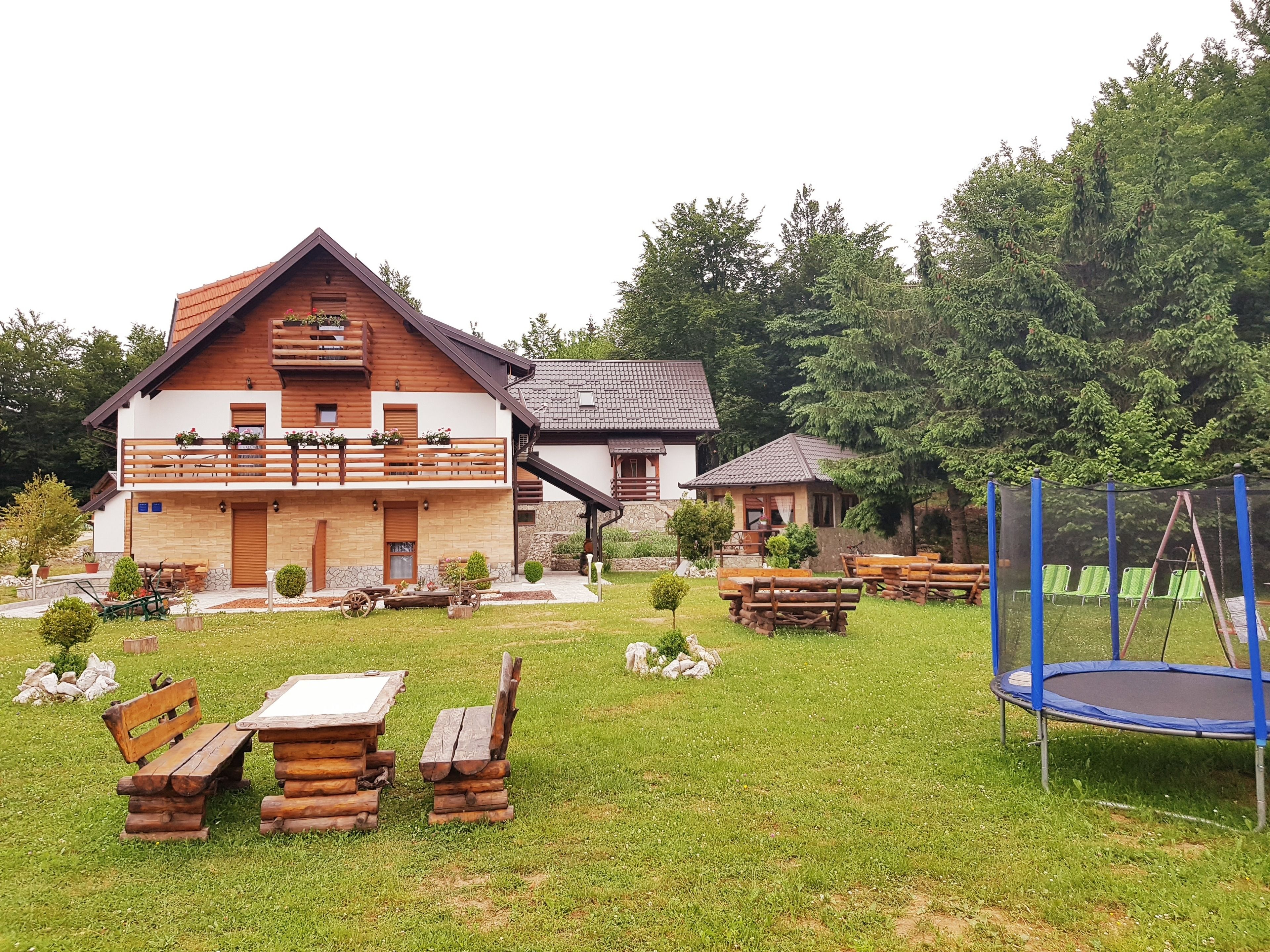 Ein rustikales Blockhaus mit Holzfassade, einem Balkon mit Blumenkästen und einem gepflegten Rasen mit Picknicktischen und einem Trampolin.