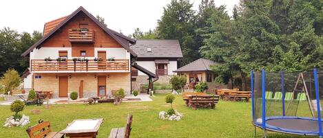Ein rustikales Blockhaus mit Holzfassade, einem Balkon mit Blumenkästen und einem gepflegten Rasen mit Picknicktischen und einem Trampolin.