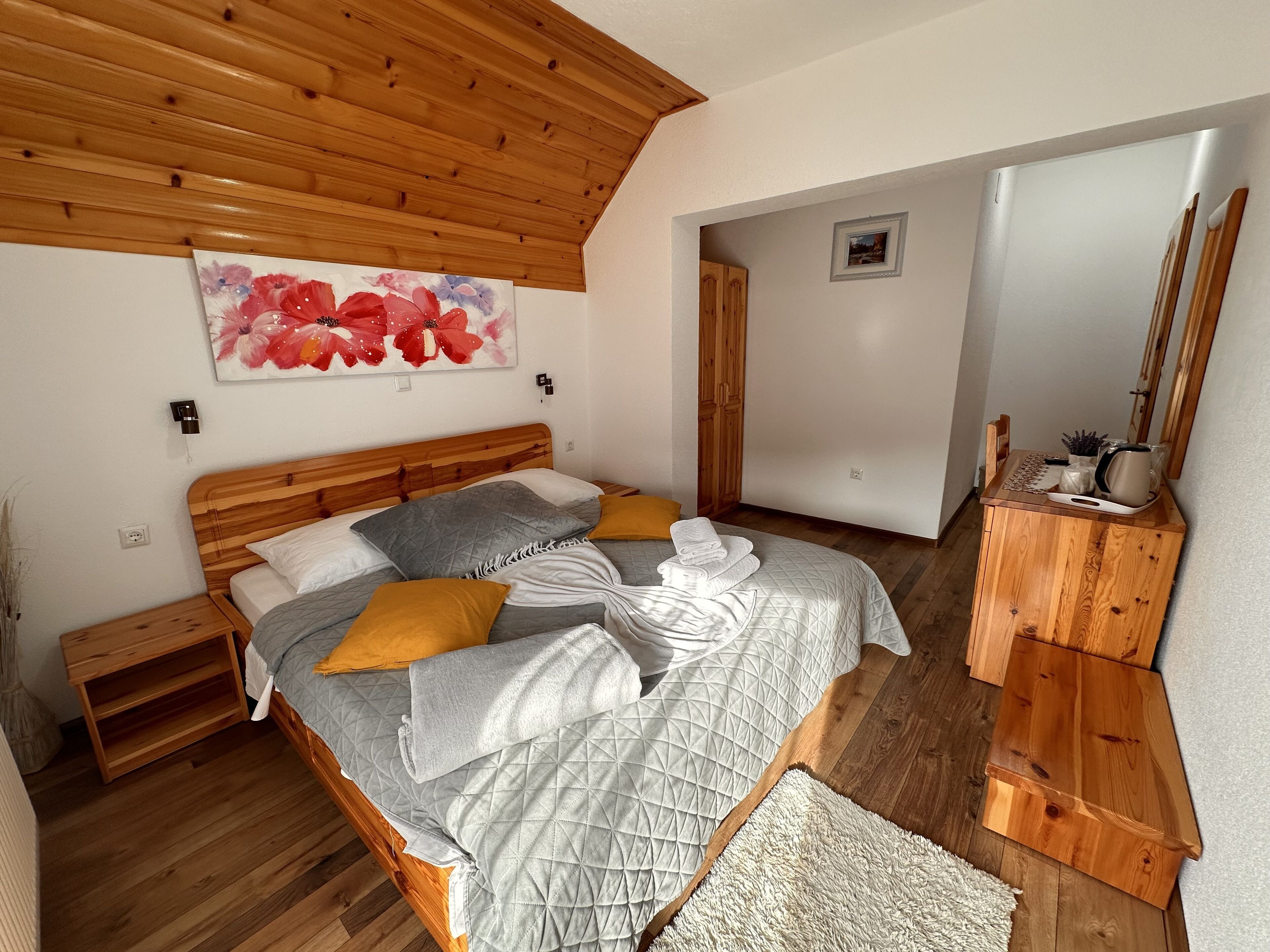 A bedroom with a wooden ceiling, a bed with multiple pillows, a bedside table, a wooden dresser, and a painting on the wall.