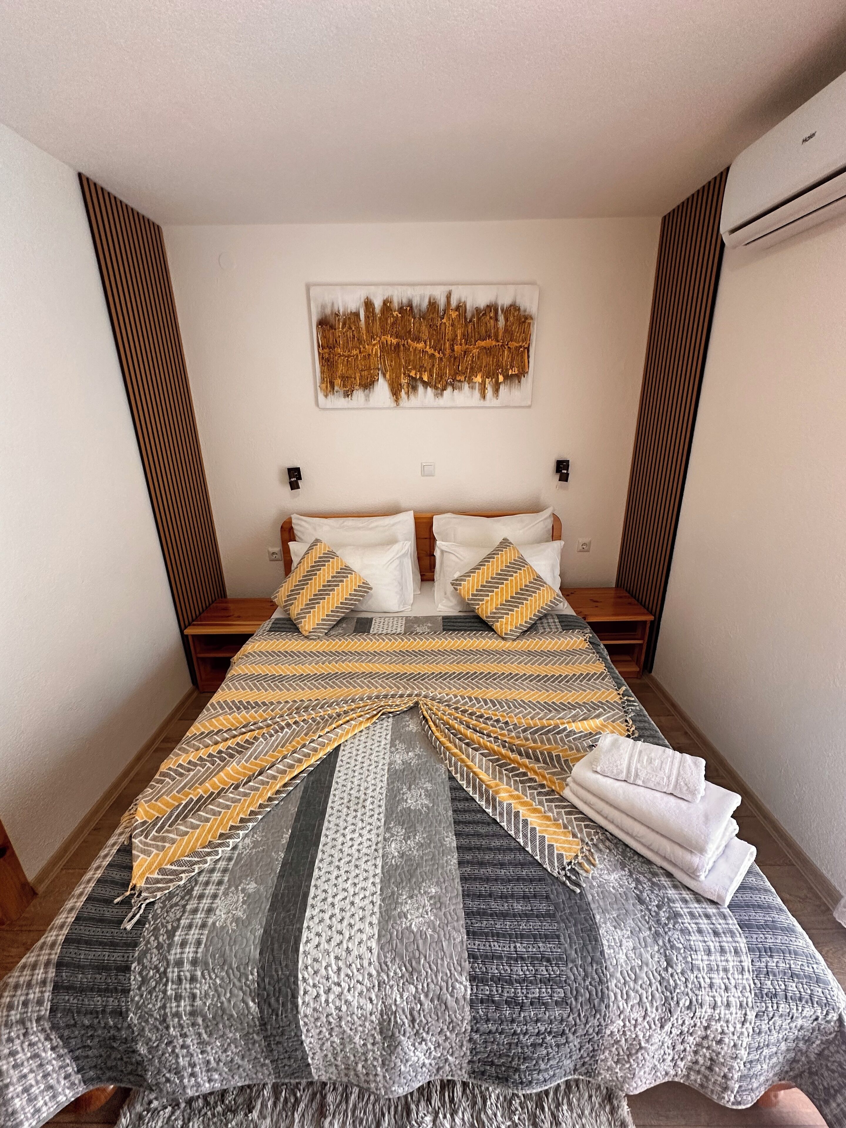 A neatly made bed with patterned bedding, a wall-mounted artwork, and a modern air conditioning unit.