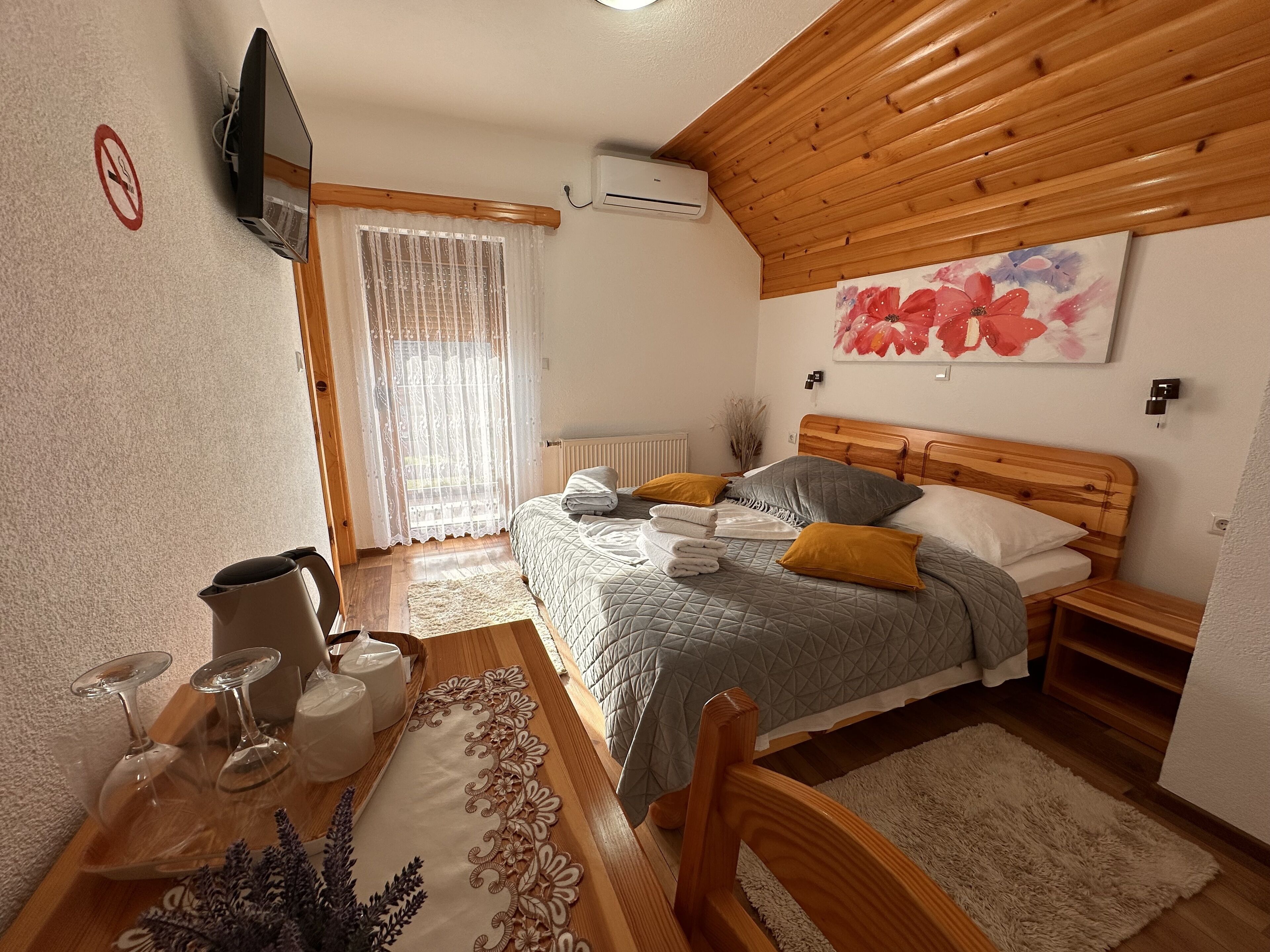 Een slaapkamer met een houten plafond, een bed met kussens, een nachtkastje, een raam met gordijnen, een aan de muur bevestigde televisie en een radiator.