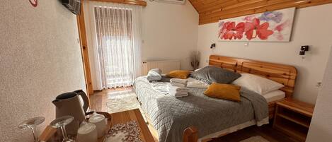 Een slaapkamer met een houten plafond, een bed met kussens, een nachtkastje, een raam met gordijnen, een aan de muur bevestigde televisie en een radiator.