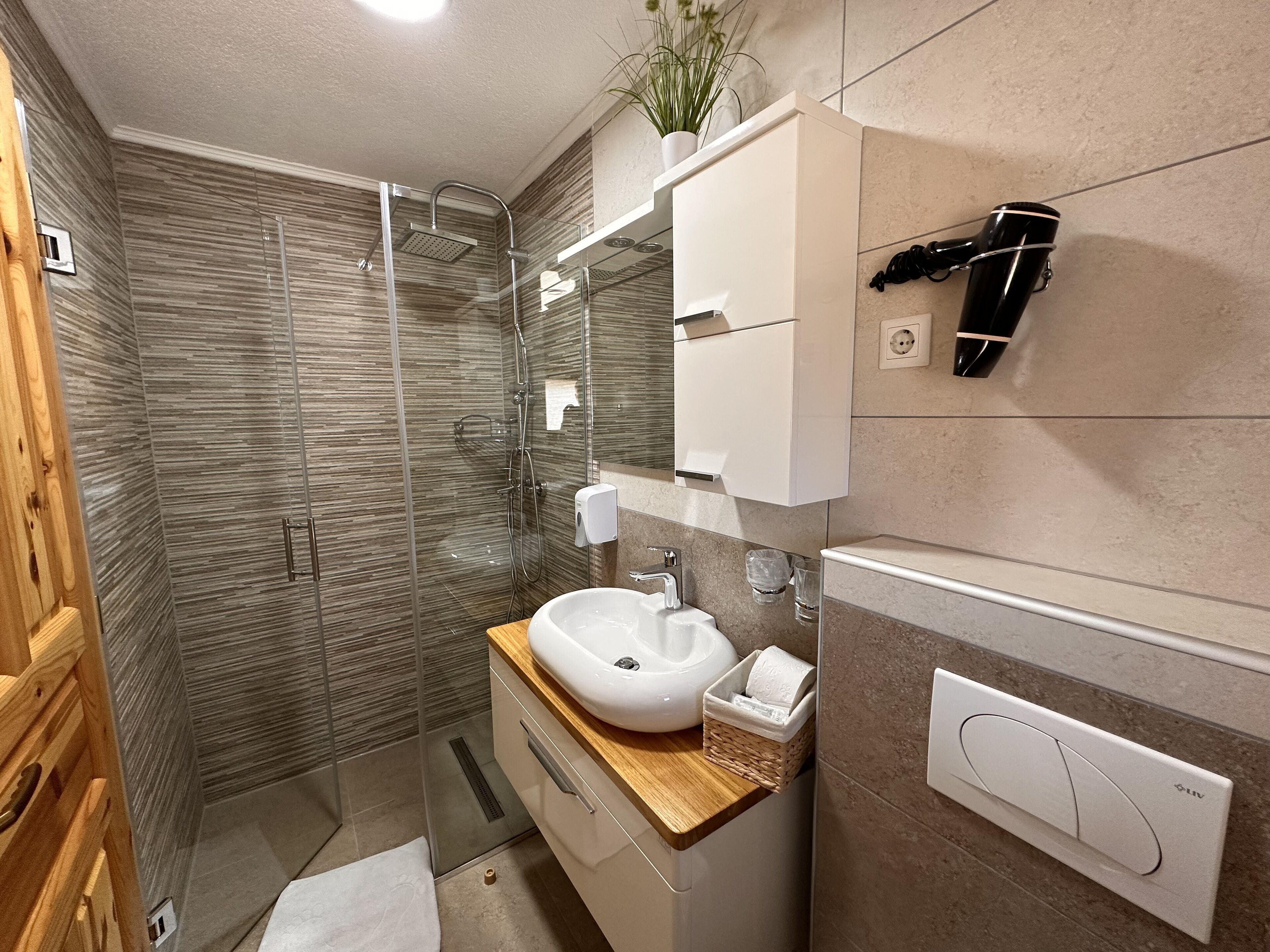 Un baño moderno con ducha envuelta en vidrio, tocador de madera, lavabo blanco y secador de pelo montado en la pared.